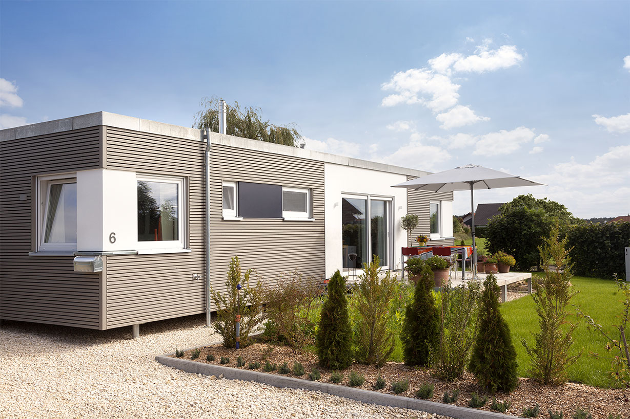 Schwoerer_FlyingSpace_Kohler_0968 Das einstöckige, moderne Minihaus verfügt über große Fenster, eine gemütliche Terrasse mit Sonnenschirm und liegt eingebettet in einen gepflegten Garten. Ein Steinpfad schlängelt sich unter dem klaren blauen Himmel durch kleine Bäume.