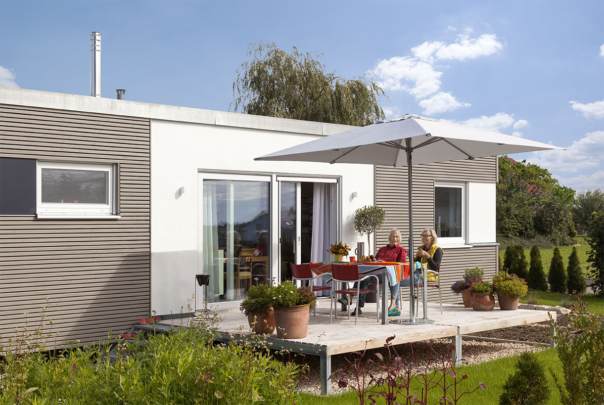 Schwoerer_FlyingSpace_Kohler_1002 Ein modernes, einstöckiges Minihaus mit Holzterrasse. Zwei Personen sitzen an einem Tisch unter einem Sonnenschirm, umgeben von Topfpflanzen.