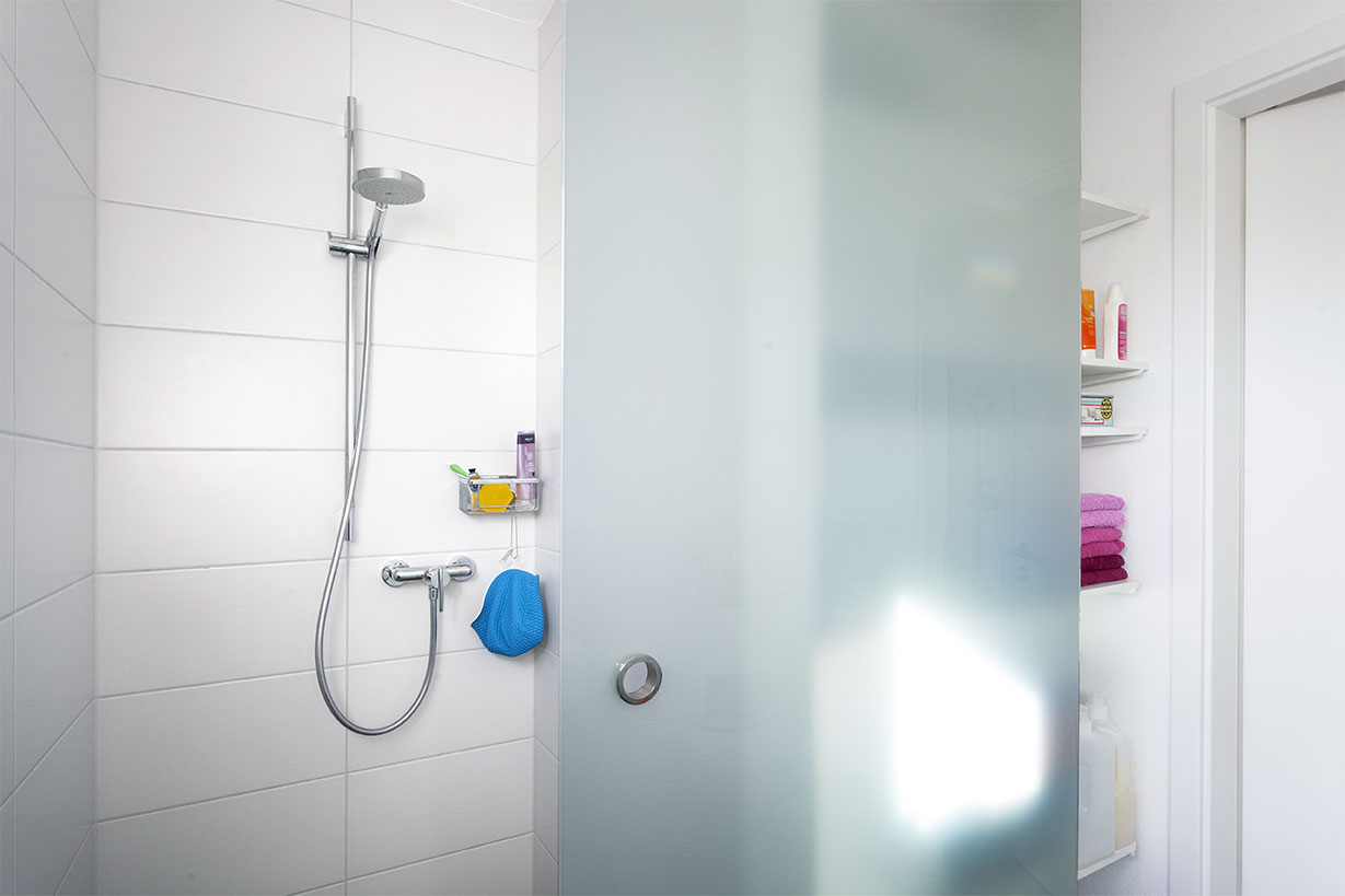 Schwoerer_FlyingSpace_Kohler_1310 Die moderne Dusche verfügt über weiß geflieste Wände, einen Handbrausenkopf und eine Milchglastür. Im Hintergrund liegen ordentlich angeordnete Handtücher und Toilettenartikel auf Regalen.