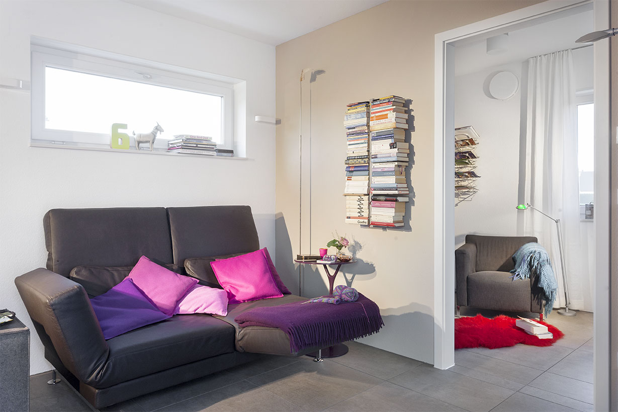 Schwoerer_FlyingSpace_Kohler_1542 Ein modernes Minihaus-Wohnzimmer mit einem schwarzen Sofa, leuchtend violetten und rosa Kissen, einem an der Wand hängenden Bücherstapel, einem Altarsessel in der Ecke und einem auffälligen roten Teppich auf dem Boden.