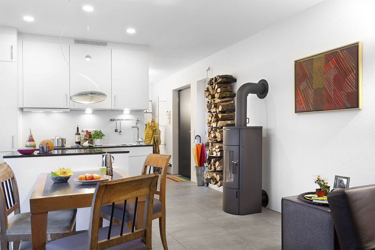 Schwoerer_FlyingSpace_Kohler_1614 Das Minihaus Alter verfügt über eine moderne offene Küche und einen Essbereich mit einem Holztisch, einem Stapel Brennholz und einem eleganten schwarzen Holzofen. Ein farbenfrohes Gemälde belebt die weiße Wand und schafft eine gemütliche und doch moderne Atmosphäre.