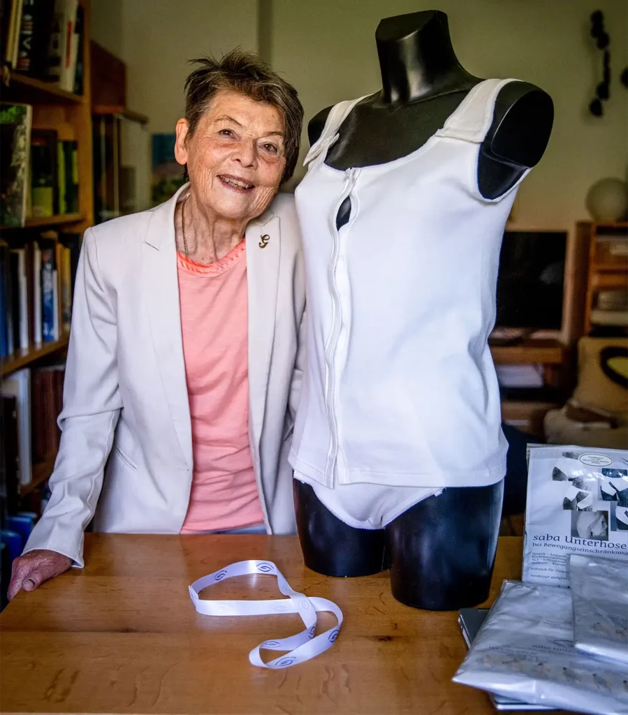 Eine ältere Frau steht neben einer schwarzen Schaufensterpuppe, die in weißer medizinischer Kleidung auf einem Tisch steht