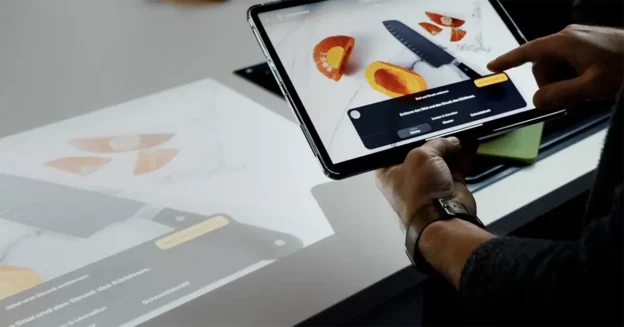 Person, die ein Tablet benutzt, um ein digitales Bild eines Messers und aufgeschnittener Früchte auf einem Tisch anzuzeigen und damit zu interagieren.
