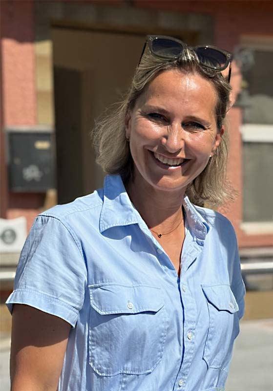 Frau in hellblauem, kurzärmeligem Hemd lächelt im Freien und trägt eine Sonnenbrille auf dem Kopf.