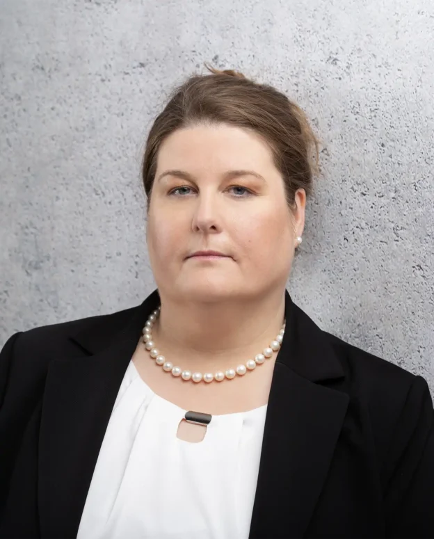 Frau in schwarzem Blazer und weißer Bluse, die eine Perlenkette trägt, posiert vor einer hellgrauen, strukturierten Wand.