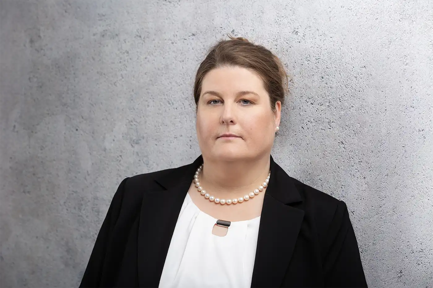 Eine Person, die einen schwarzen Blazer, eine weiße Bluse und eine Perlenkette trägt, steht vor einer hellgrauen, strukturierten Wand.