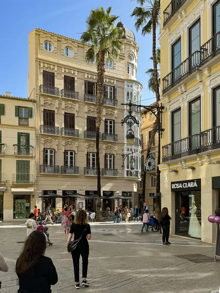 Die Menschen spazieren über einen sonnenbeschienenen Stadtplatz, der von Palmen und historischen Gebäuden gesäumt ist, darunter Geschäfte und ein Brautmodengeschäft.