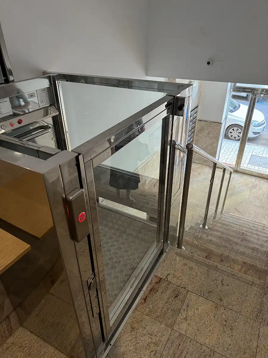 Ein kleiner gläserner Aufzug für Barrierefreiheit ist neben einer Treppe in einem Gebäudeeingangsbereich installiert.