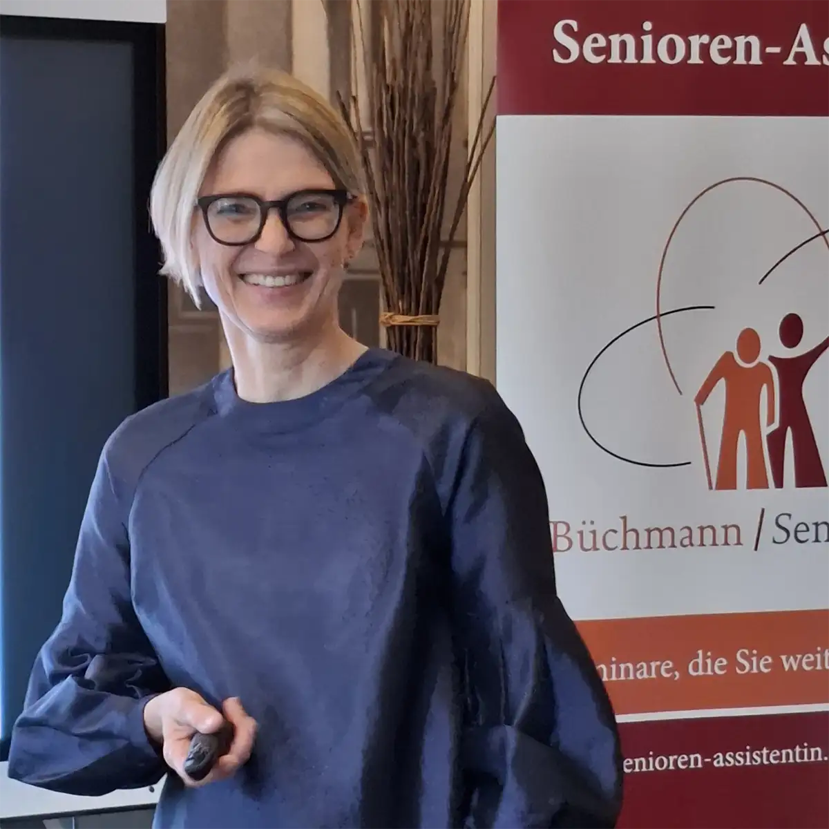 Eine lächelnde Seniorenassistentin mit kurzen blonden Haaren und Brille steht vor einem Senioren-Assistenz-Seminarbanner.
