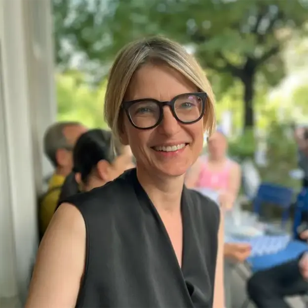 Seniorenassistentin mit kurzen blonden Haaren und Brille lächelnd im Freien, Menschen und Grün im Hintergrund.