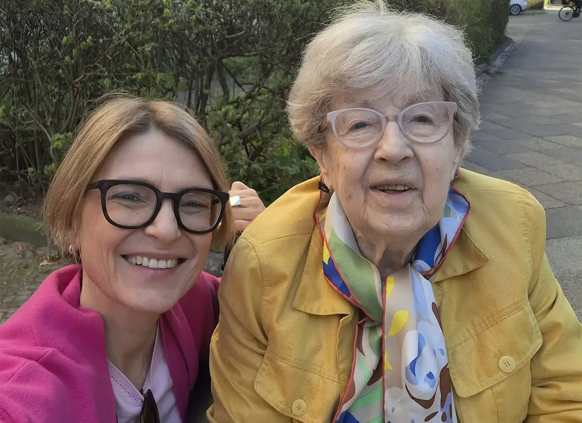 Zwei Frauen, eine davon eine Seniorenassistentin, lächeln im Freien, im Hintergrund eine Grünanlage und ein Bürgersteig.