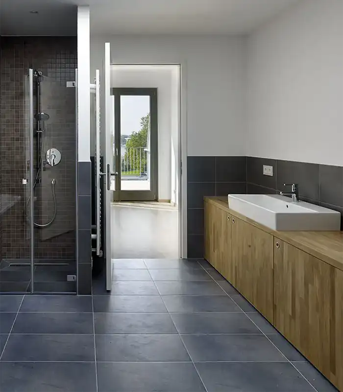Modernes Badezimmer mit Glasdusche, großem Waschbecken, Holzschränken und einer Tür, die zu einem hellen Flur führt.