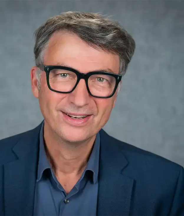 Ein Mann mittleren Alters mit Brille und grauem Haar trägt einen blauen Blazer und ein blaues Hemd und lächelt in die Kamera.