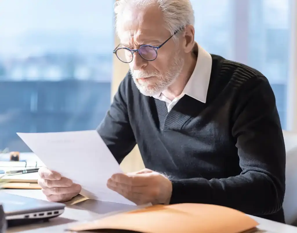 Älterer Mann mit Brille liest ein Dokument an einem Schreibtisch mit Papieren, Laptop und einem Fenster im Hintergrund.