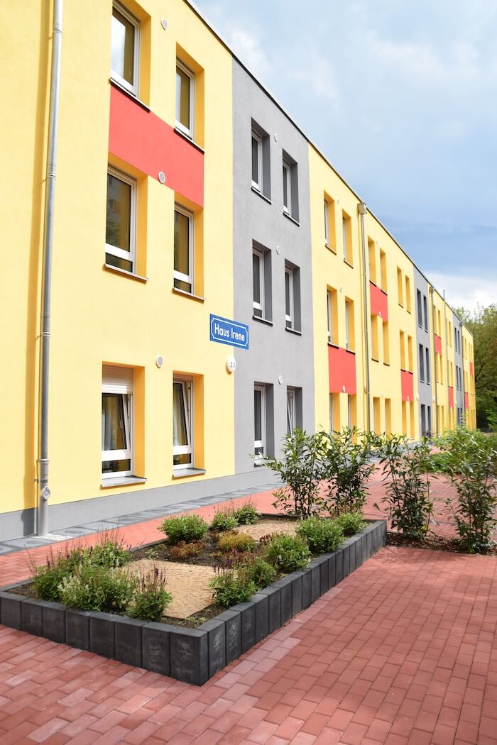 Dreistöckiges Wohnhaus mit gelben und grauen Wänden, roten Akzenten und einem kleinen Vorgarten.