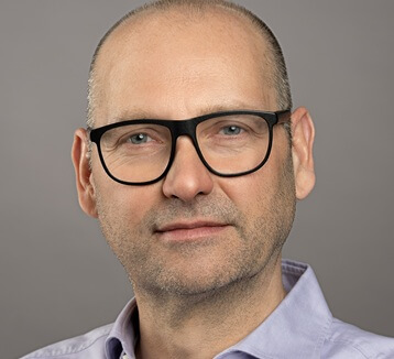 Ein Mann mittleren Alters mit Brille, der ein hellviolettes Hemd trägt, posiert vor einem schlichten grauen Hintergrund.