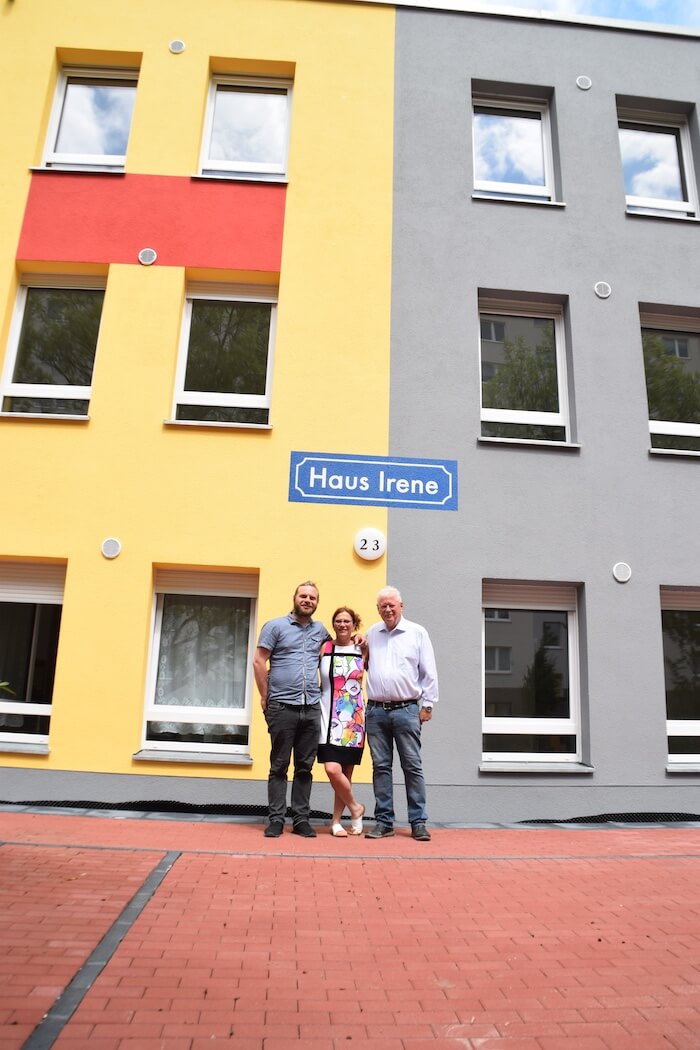 Drei Personen stehen vor einem Gebäude mit der Aufschrift Haus Irene und gelben und grauen Außenwänden.