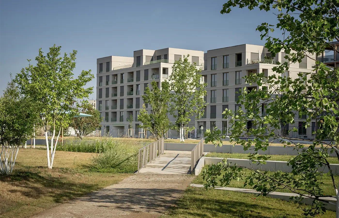 ANUNDO_Gemeinschaft-Projekt-AnsichtvomPark Modernes Mehrfamilienhaus mit Balkonen, umgeben von Bäumen und einer gepflegten Grünfläche unter freiem Himmel.