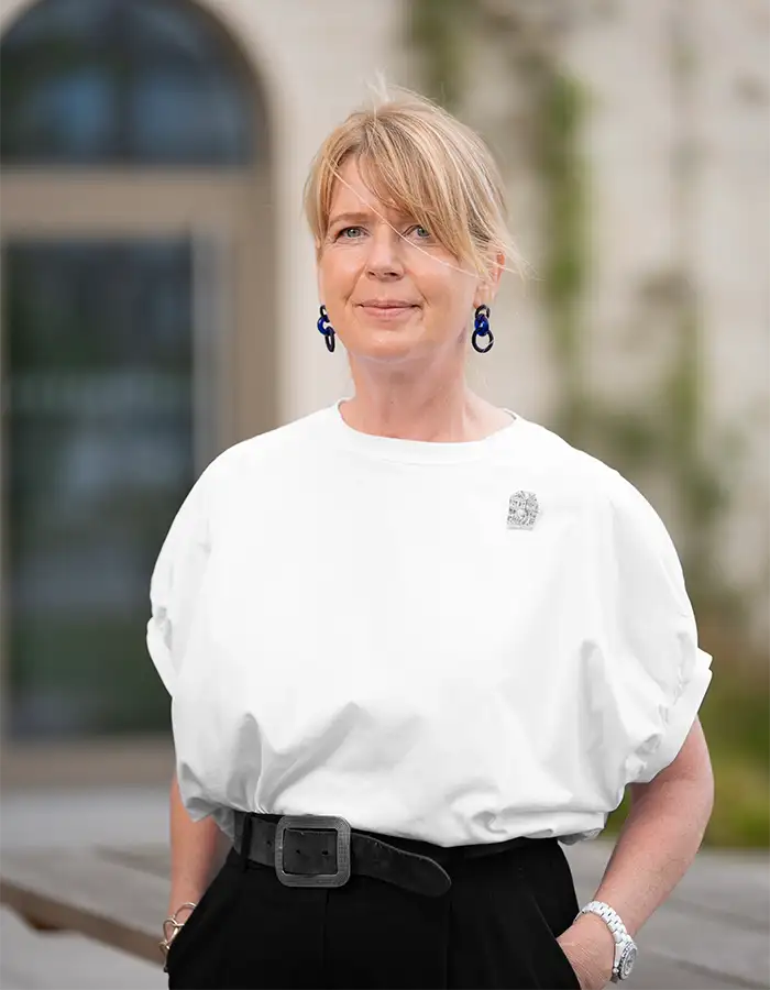 Eine Frau mit blondem Haar, weißem Hemd und schwarzer Hose steht draußen vor einem verschwommenen Gebäude.