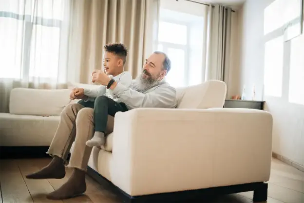 Ein älterer Mann und ein kleiner Junge sitzen zusammen auf einer weißen Couch in einem hellen Wohnzimmer, beide lächeln.