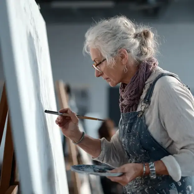 Ältere Frau mit Brille und Latzhose malt in einem gut beleuchteten Atelier auf einer Leinwand.