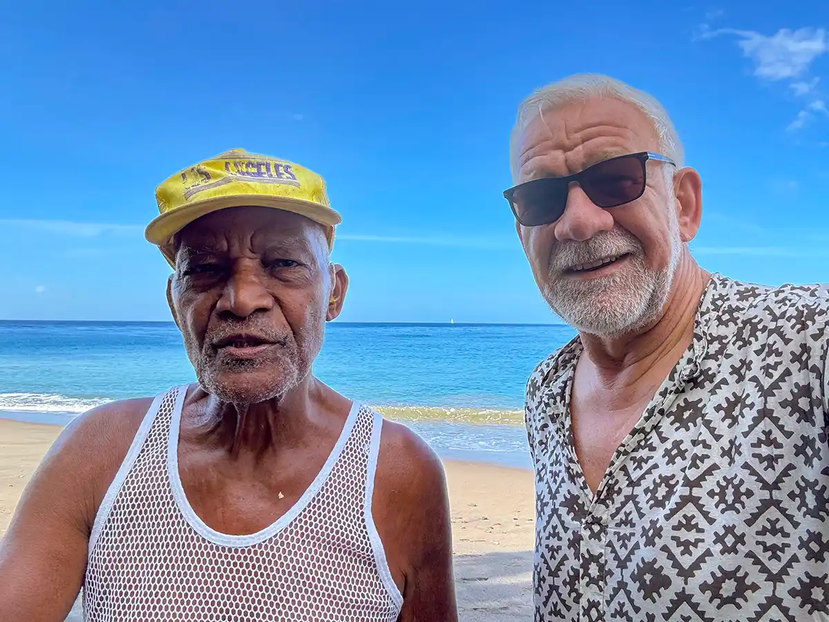 Longevity-Martinique Zwei ältere Männer stehen an einem Strand mit blauem Himmel und Meer im Hintergrund und lächeln in die Kamera.