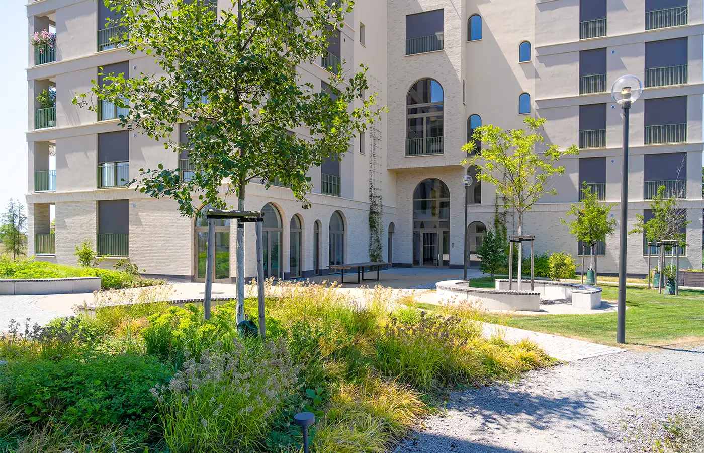 Wohnen-55plus-Gemeinschaftsgarten-ANUNDO Modernes Wohnhaus mit großen Fenstern, umgeben von Grünanlagen und einem kleinen Innenhof.