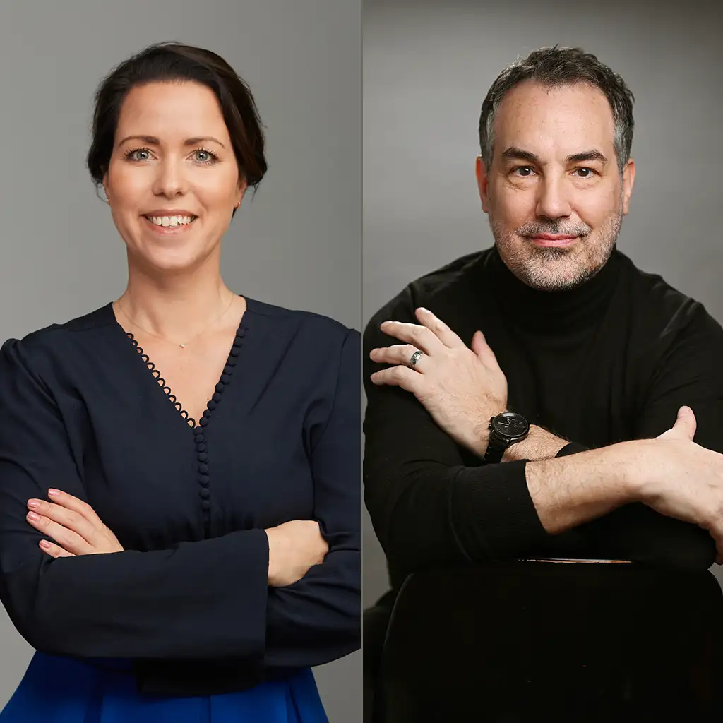 Eine Frau in einer marineblauen Bluse und ein Mann in einem schwarzen Rollkragenpullover sitzen mit verschränkten Armen vor neutralen Hintergründen.