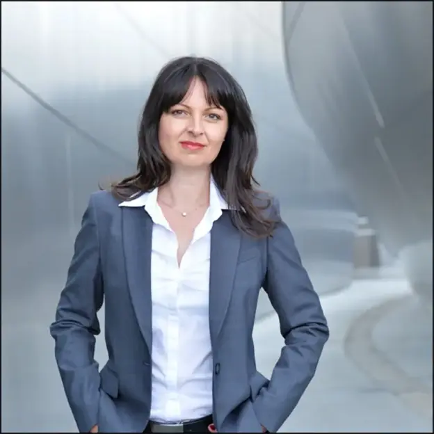 Eine Frau in grauem Blazer und weißem Hemd steht im Freien vor einer modernen Metallstruktur und lächelt.