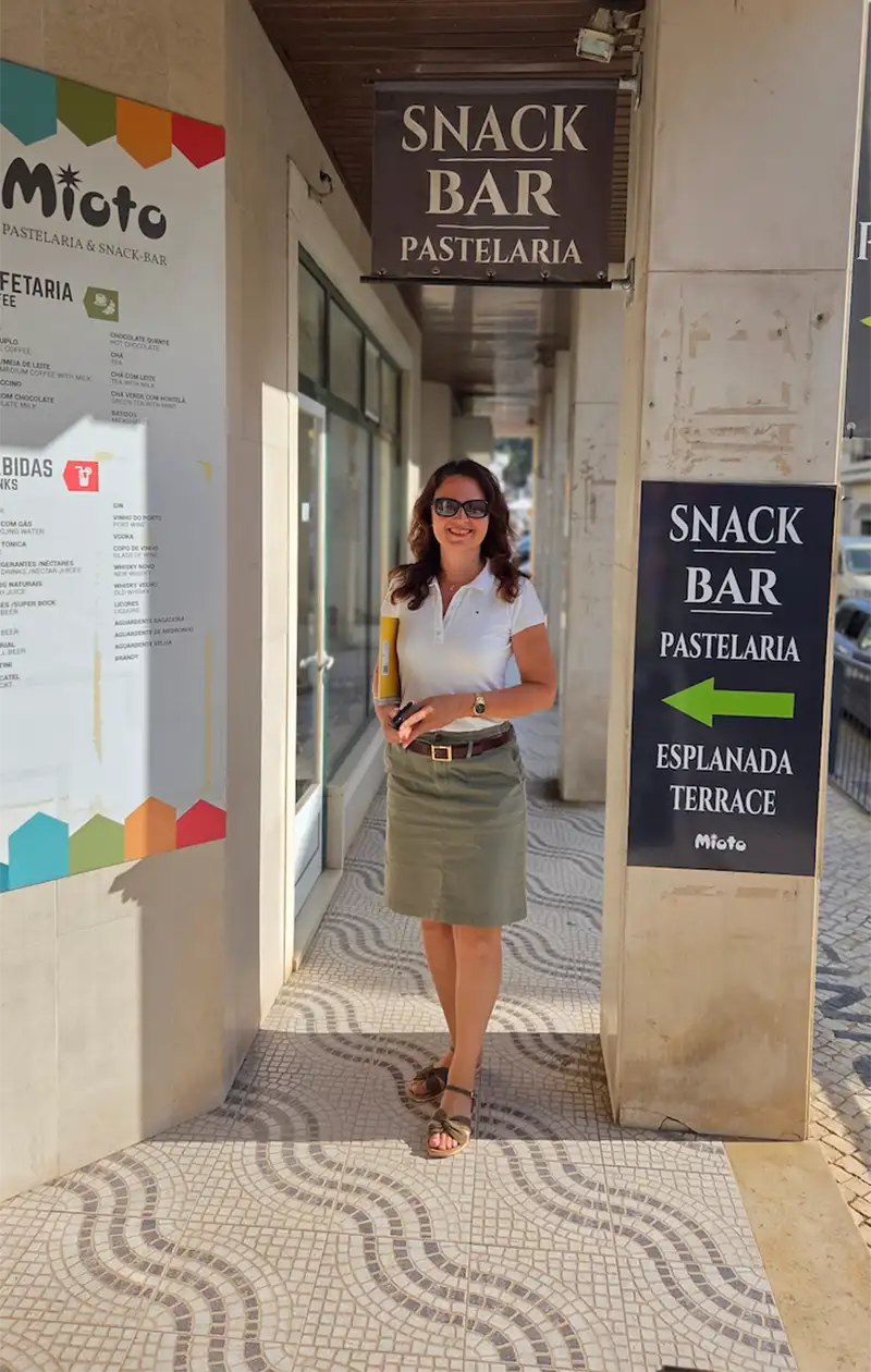 Jana-Mrowetz-Wohnkonzept-Urban-Cell-Portugal Eine Frau mit Sonnenbrille steht vor einem Imbiss und einer Pasteleria auf einem gekachelten Bürgersteig im Tageslicht.