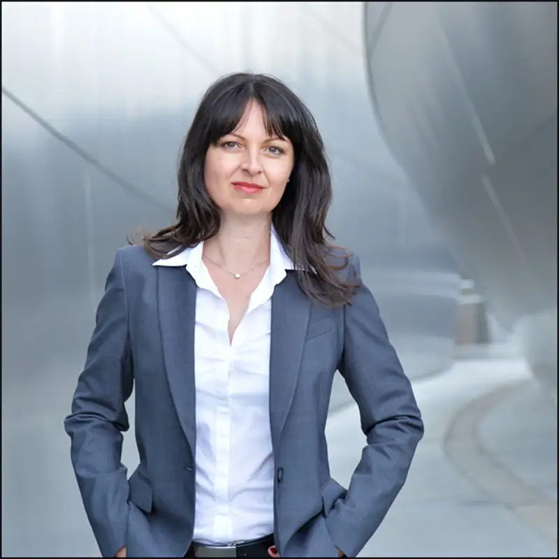 Eine Frau in grauem Blazer und weißem Hemd steht im Freien vor einer modernen Metallstruktur und lächelt.