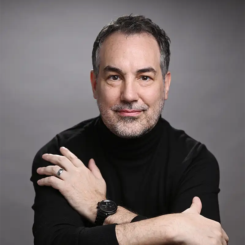 Ein Mann mit kurzen dunklen Haaren und einem Bart, der einen schwarzen Rollkragenpullover trägt, posiert mit verschränkten Armen vor einem grauen Hintergrund.