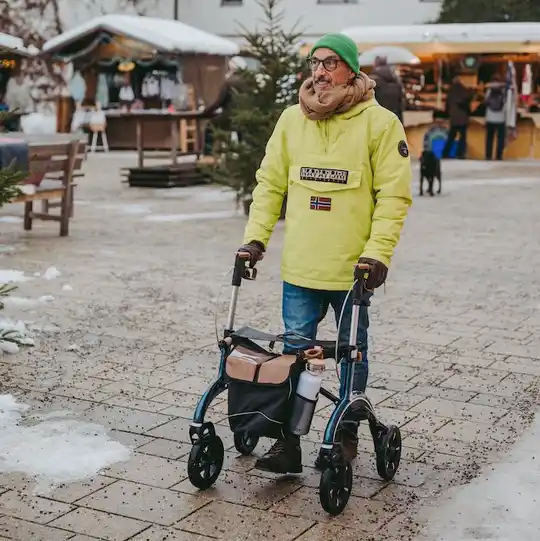 Eine Person mit einer grünen Mütze benutzt einen Rollator auf einem verschneiten Weg in der Nähe von Marktständen.