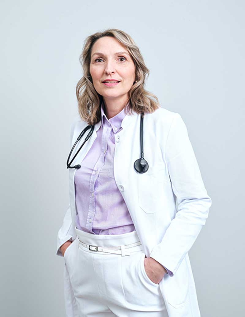 Sandra-Eifert Eine Frau in weißem Laborkittel und Stethoskop steht, weibliche Langlebigkeit verkörpernd, vor einem schlichten Hintergrund.