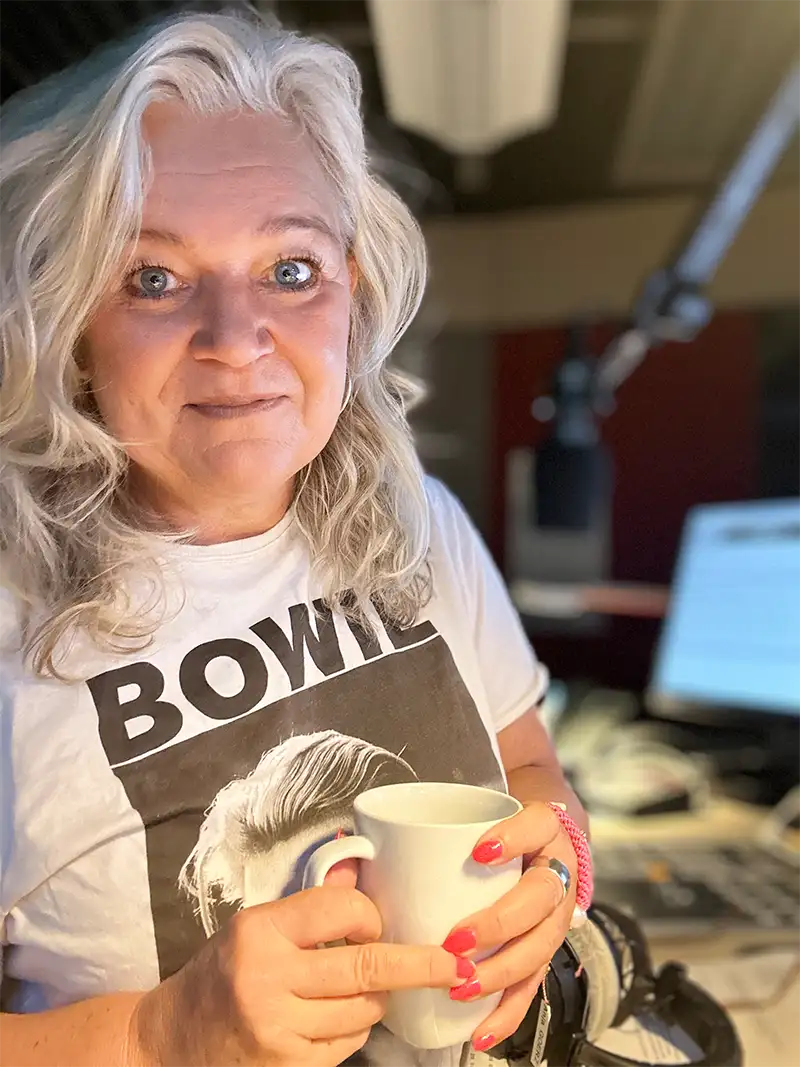 Eine Frau mit grauen Haaren und einem Bowie-Shirt steht für ein Interview am Mikrofon und hält einen Becher im Stil von Anja Goerz.