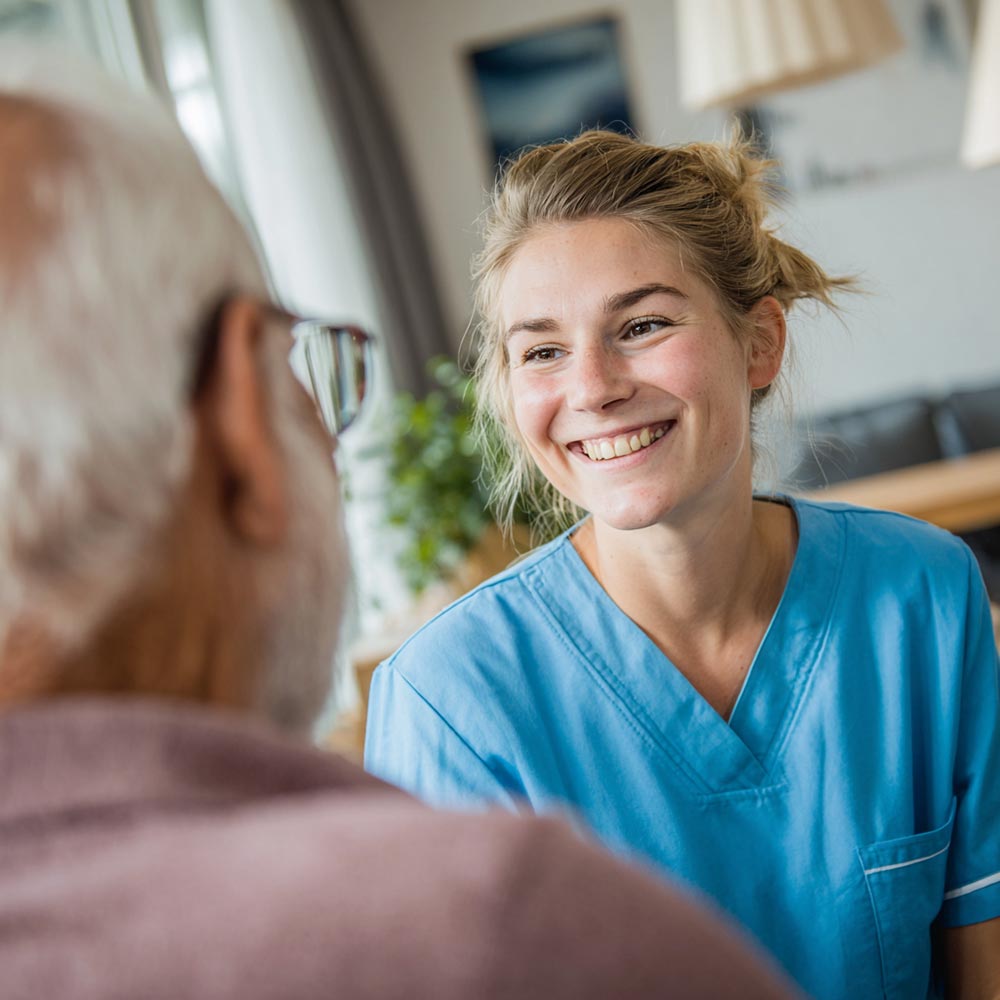 Eine Pflegekraft aus dem Ausland lächelt in blauen Kittel, während sie mit einem älteren Herrn spricht.