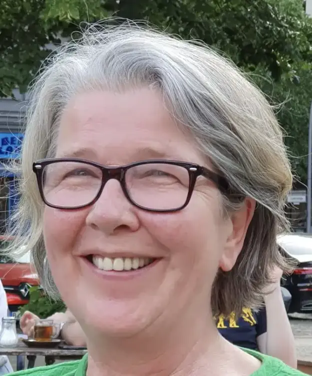 Dr. Bettina Held, mit kurzen grauen Haaren und Brille, lächelt im Freien - Bäume und Autos im Hintergrund.