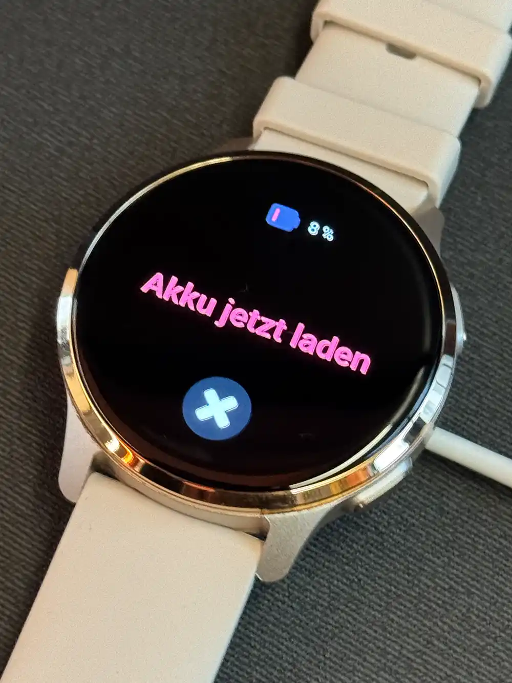 Patronus-Notfalluhr-Akkulaufzeit Eine Patronus Notfalluhr zeigt "Akku jetzt laden" mit einem Symbol für einen niedrigen Batteriestand und einem weißen Band an, das auf einer Stoffoberfläche liegt.