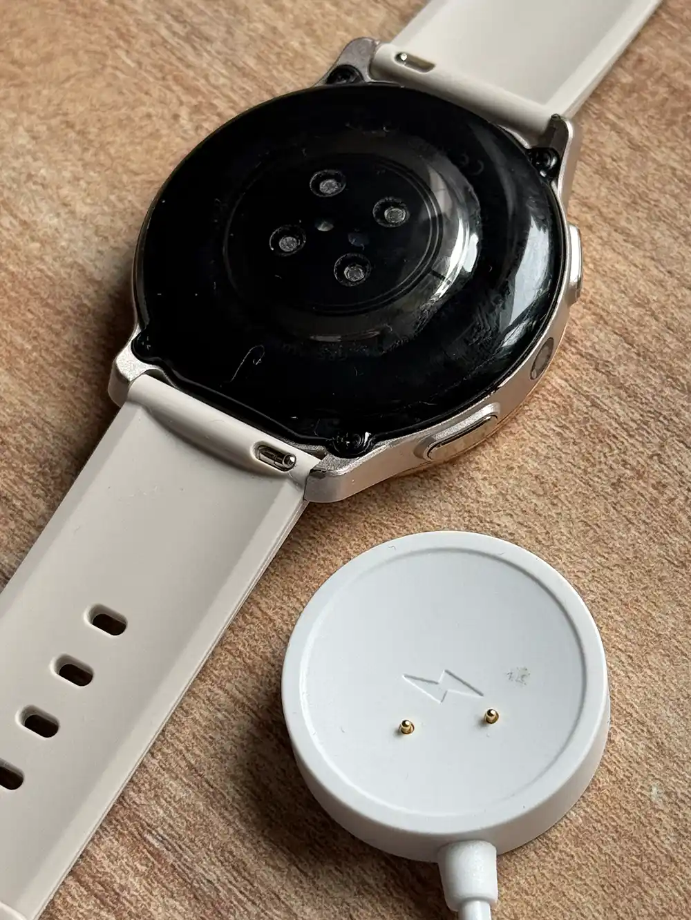 Patronus-Notfalluhr-Test-Laden Eine Smartwatch mit weißem Armband liegt mit der Vorderseite nach unten neben einer runden weißen magnetischen Ladestation auf einer Holzoberfläche.