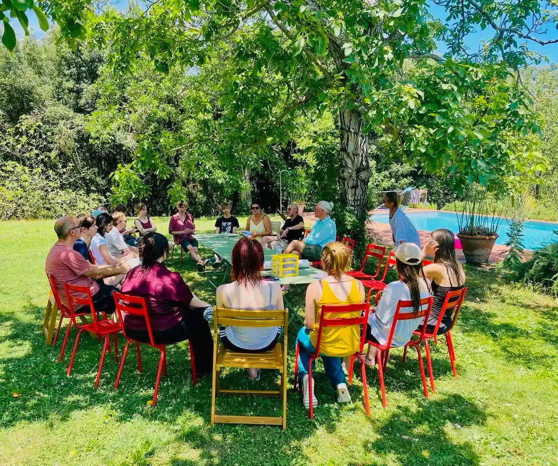 Wohndorf-Borgo-Batone-Pool Eine Gruppe von Menschen sitzt im Kreis auf roten Stühlen im Freien unter einem großen Baum in der Nähe eines Swimmingpools.