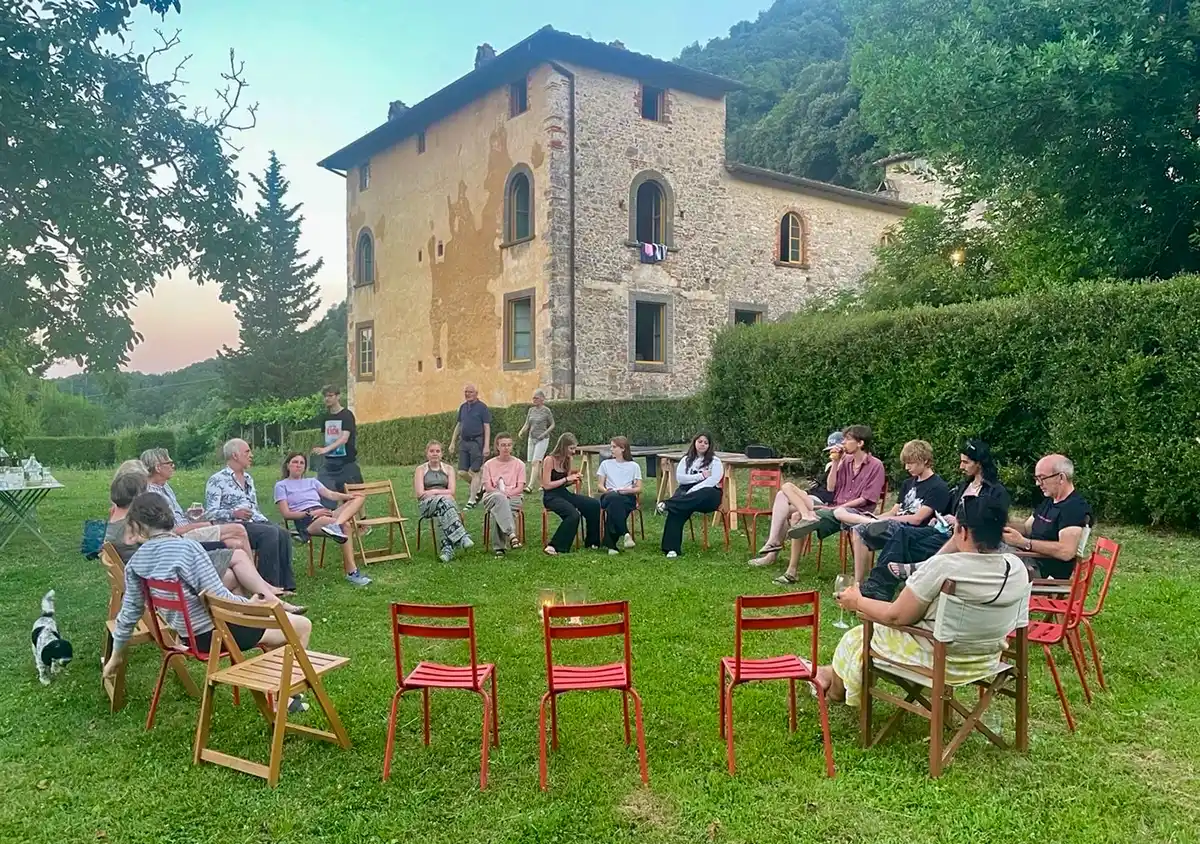 Wohndorf-Borgo-Batone-Toskana Eine Gruppe von Menschen sitzt in einem Stuhlkreis auf einem Rasen vor einem steinernen Gebäude, mit Bäumen und Hügeln im Hintergrund.