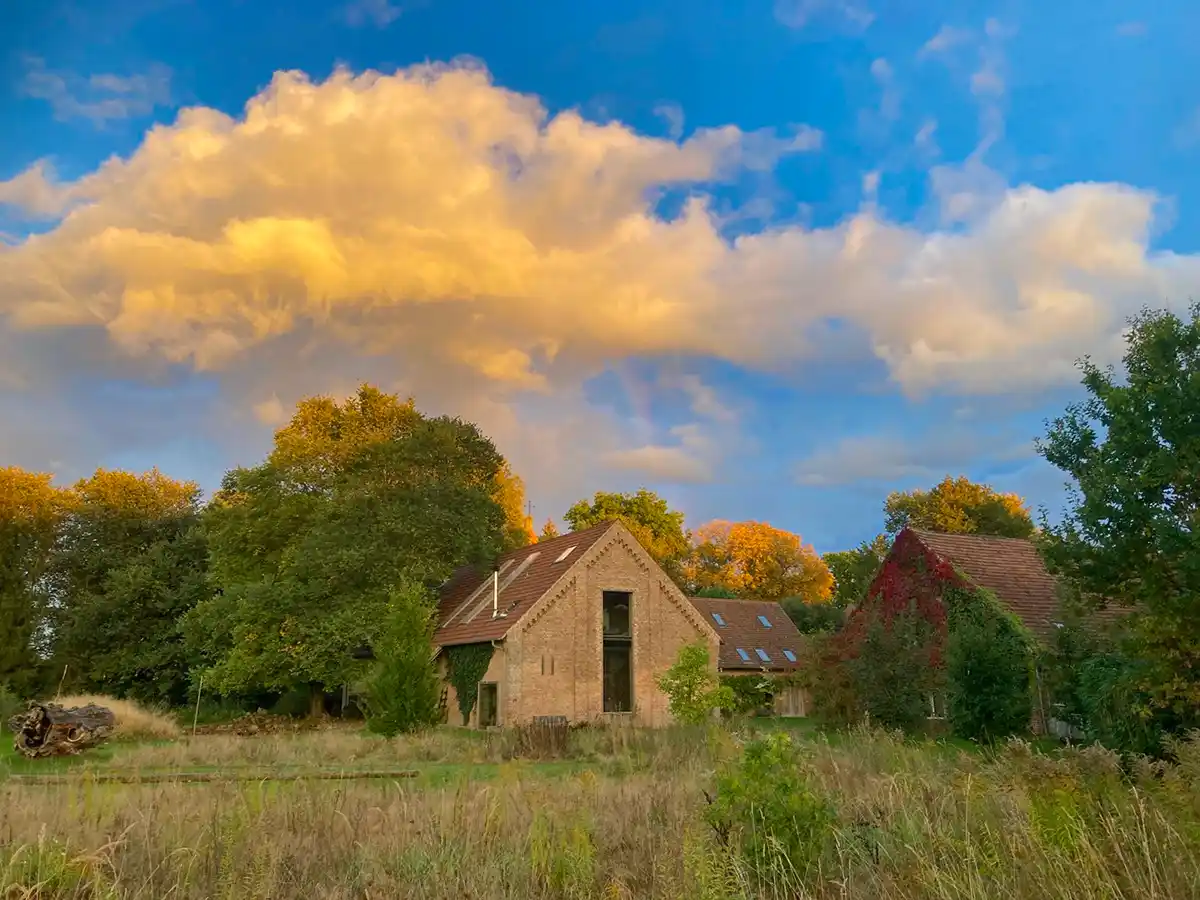 Wohndorf-Re-hof-Abend Ein Backsteinhaus mit Schrägdach steht auf einer Wiese, umgeben von Bäumen und unter einem teilweise bewölkten Himmel.