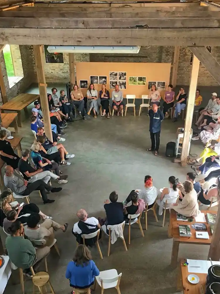 Wohndorf-Re-hof-Besprechung Eine Gruppe von Menschen, die in einem großen Kreis sitzen, hört einer Person zu, die in der Mitte eines rustikalen, offenen Raums spricht.