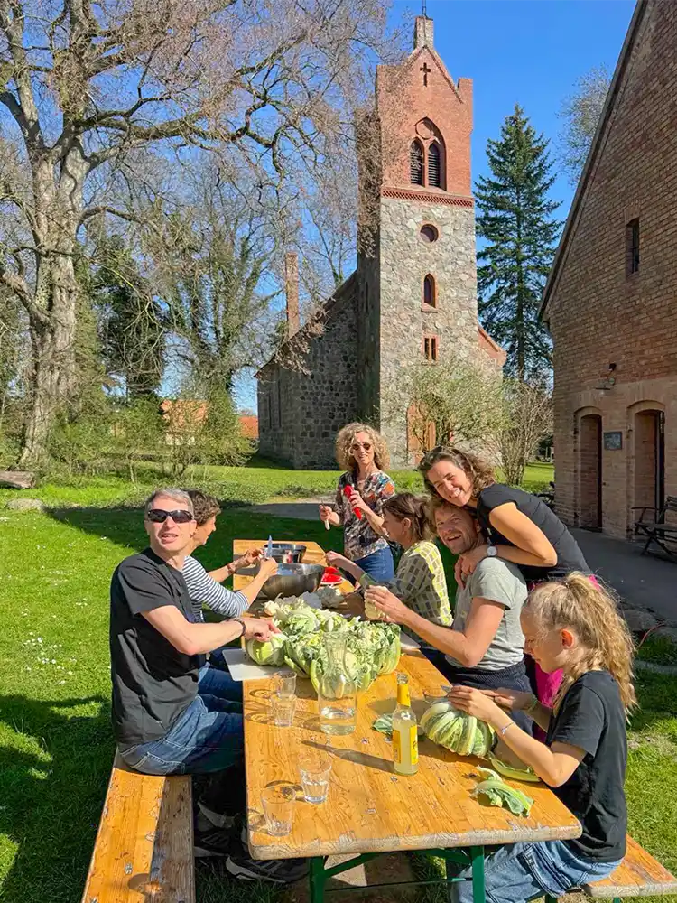 An einem sonnigen Tag sitzt und steht eine Gruppe von Menschen um einen Picknicktisch in der Nähe einer alten Steinkirche.