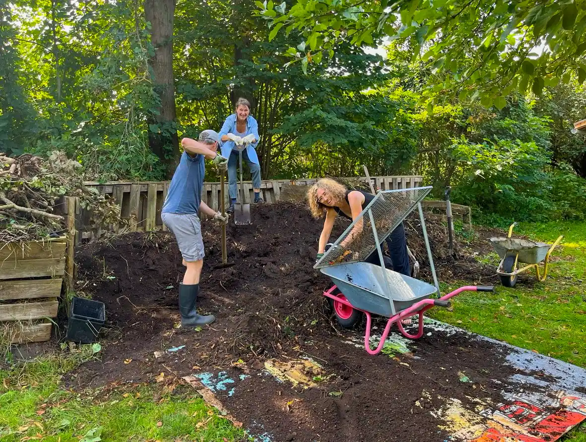 Wohndorf-Re-hof-Gartenarbeit Drei Personen arbeiten in einem Garten und schaufeln Erde in eine Schubkarre neben Komposttonnen und Bäumen.