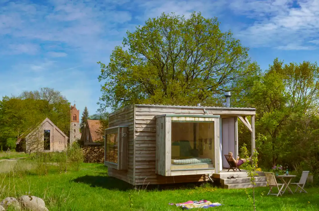 Wohndorf-Re-hof-Sommer Eine kleine, moderne Holzhütte mit großen Fenstern steht auf einer Wiese, umgeben von Bäumen und Gartenmöbeln.