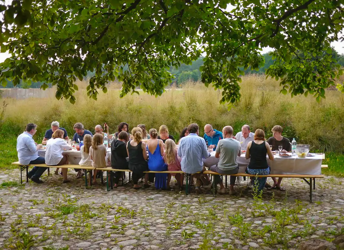Wohndorf-Re-hof-Tafel Eine Gruppe von Menschen sitzt an einem langen Tisch im Freien und isst gemeinsam auf einer Kopfsteinpflaster-Terrasse unter einem Baum.