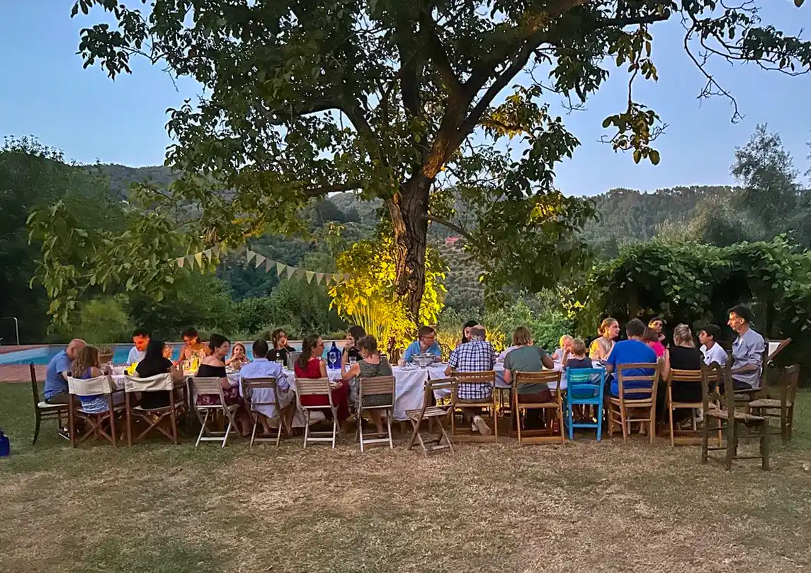 bzw-Dorf-Gemeinschaft Eine Gruppe von Menschen sitzt um einen langen Tisch im Freien unter einem Baum und isst gemeinsam zu Abend.