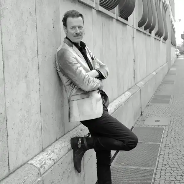 Ein Mann im Blazer lehnt mit verschränkten Armen und einem angewinkelten Bein an einer Steinmauer und verkörpert damit das Rollenmodell Alter.