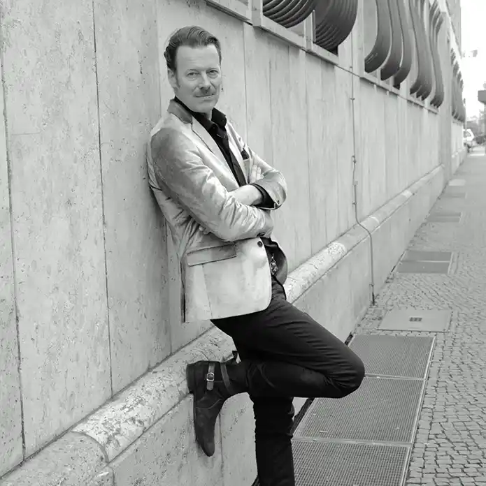 Ein Mann im Blazer lehnt mit verschränkten Armen und einem angewinkelten Bein an einer Steinmauer und verkörpert damit das Rollenmodell Alter.
